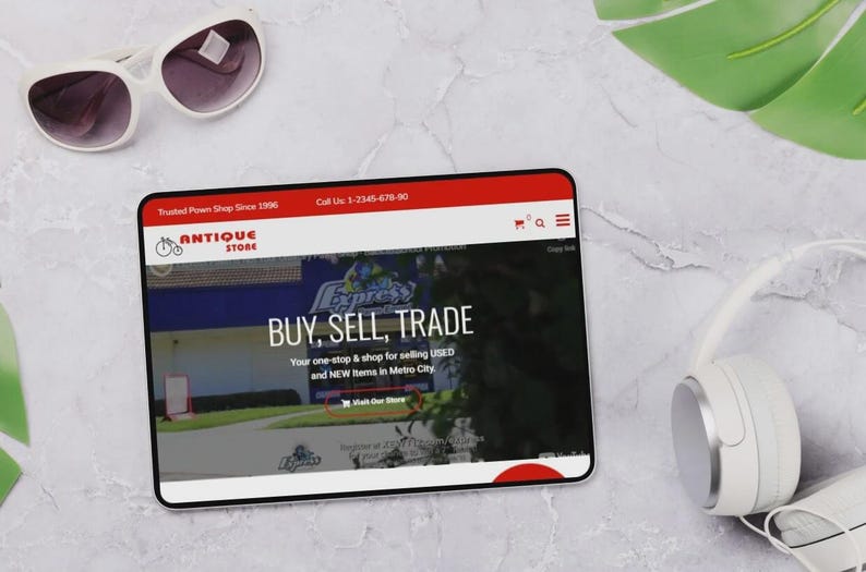 May include: A tablet showing a website with the words "BUY, SELL, TRADE" and "ANTIQUE STORE". White sunglasses and headphones are placed beside the tablet. The website also says "Trusted Pawn Shop Since 1996".