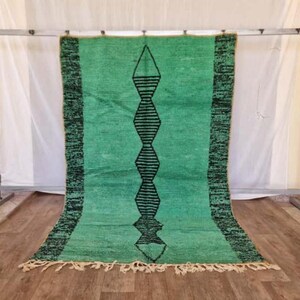 May include: A rectangular, hand-woven rug in shades of green with a central black geometric design. The rug has a fringe along the bottom edge and a dark green border. The rug is displayed against a white backdrop.
