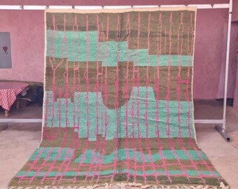 May include: A large, rectangular rug with a geometric pattern. The rug features a combination of green, brown, and pink colors. The design includes vertical and horizontal lines, creating a grid-like appearance. The rug is displayed on a metal rack.