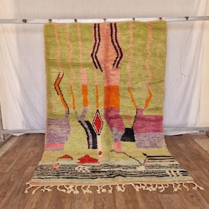 May include: A hand-woven, rectangular rug with a light green background and colorful geometric designs. The rug features vertical stripes in pink and orange, along with diamond and vase-like shapes in red, purple, and black. The bottom edge has a black and white striped pattern and a fringe.