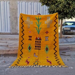 May include: A mustard yellow rug with a geometric design. The rug features a central pattern of crosses, diamonds, and other shapes in green, black, red, and blue. The edges have a zig-zag pattern. The rug is displayed outdoors.