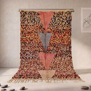May include: A rectangular area rug featuring a dense, multicolored speckled pattern of small squares. The design includes three large, vertically aligned triangles in shades of pink, gray, and cream, finished with a knotted fringe along the bottom edge.