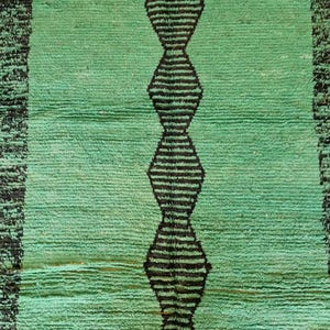 May include: A rectangular, hand-woven rug in shades of green with a central vertical design of black diamond shapes. The rug has a textured surface and a black border on each side. The rug is made of a soft material.