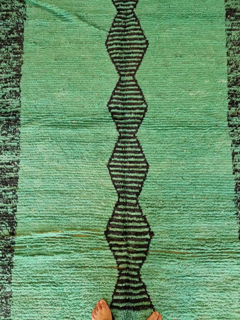 May include: A rectangular, hand-woven rug in shades of green with a central vertical design of black diamond shapes. The rug has a textured surface and a black border on each side. The rug is made of a soft material.