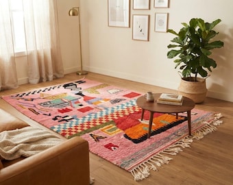May include: A rectangular area rug featuring a pink base color with a multi-colored geometric pattern including red, orange, blue, and black shapes. The rug has a textured pile, a fringed edge on the short sides, and a variety of abstract motifs.