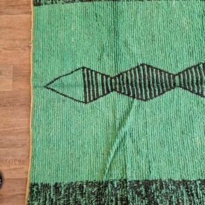May include: A rectangular, green rug with a black geometric design. The rug features a central diamond shape with vertical lines and black borders at the top and bottom. The rug is made of a textured material.