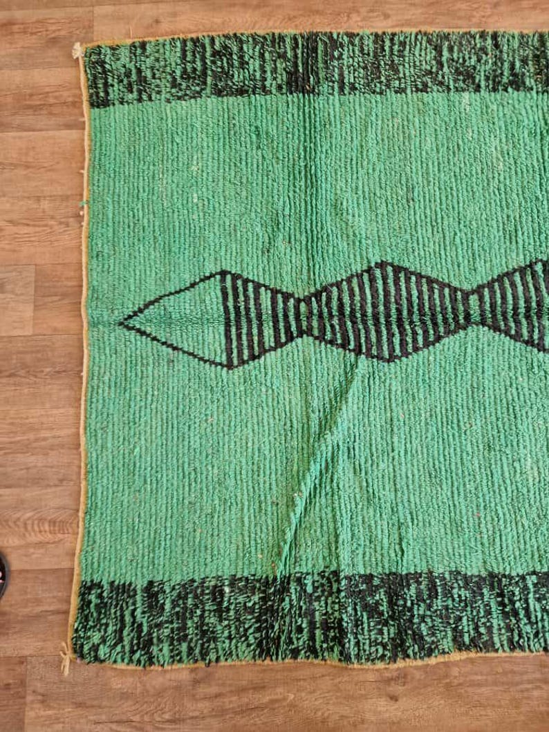 May include: A rectangular, green rug with a black geometric design. The rug features a central diamond shape with vertical lines and black borders at the top and bottom. The rug is made of a textured material.