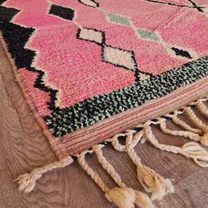 May include: A pink and black geometric patterned rug with a textured surface. The rug features diamond and rectangular shapes in black, white, and light green. The rug has a fringe of knotted tassels.