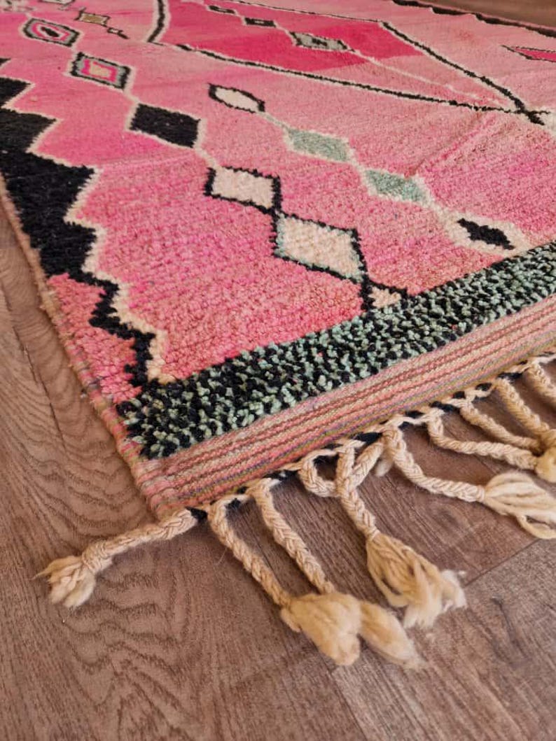 May include: A pink and black geometric patterned rug with a textured surface. The rug features diamond and rectangular shapes in black, white, and light green. The rug has a fringe of knotted tassels.