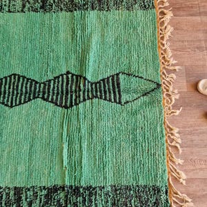 May include: A rectangular, hand-woven rug with a vibrant green field and black geometric patterns. The rug features a border of black fringe and a central design of black diamond shapes. The edges have a natural fringe.