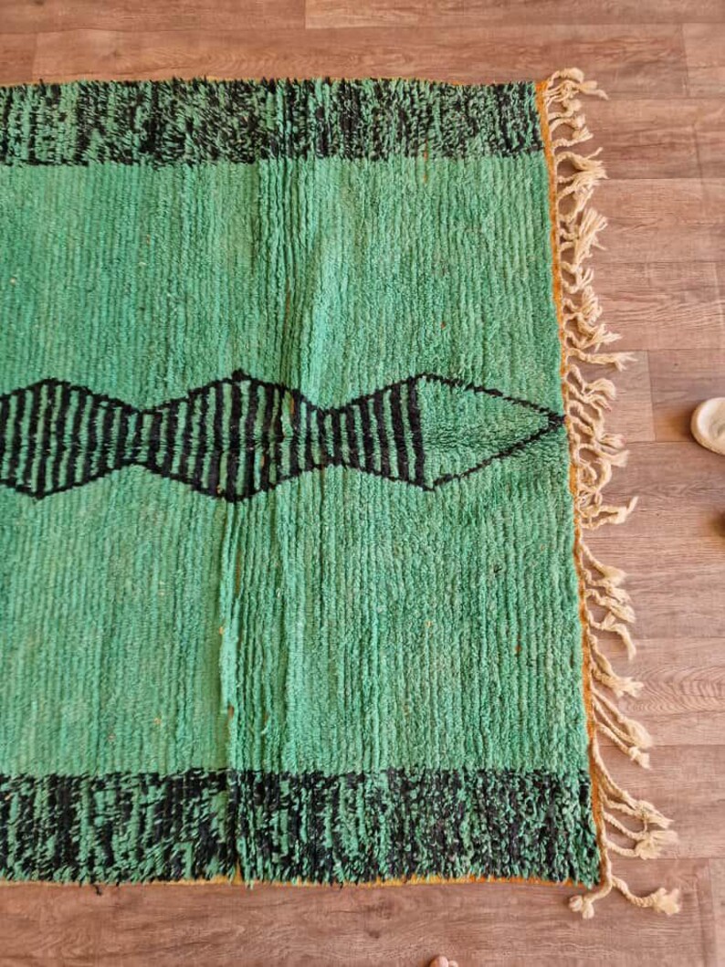 May include: A rectangular, hand-woven rug with a vibrant green field and black geometric patterns. The rug features a border of black fringe and a central design of black diamond shapes. The edges have a natural fringe.