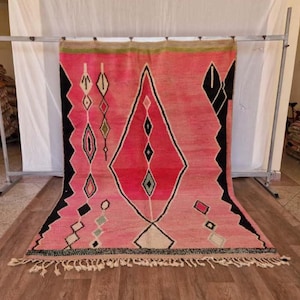 May include: A pink and red geometric patterned rug with black and white accents. The rug features a large diamond shape in the center and smaller diamond and rectangular shapes throughout. The rug has a fringe along the bottom edge.