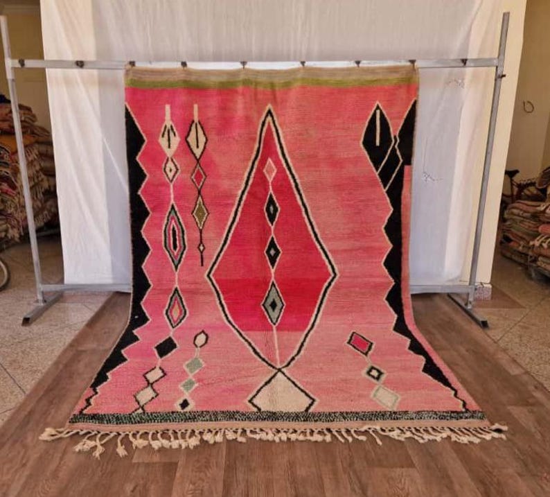 May include: A pink and red geometric patterned rug with black and white accents. The rug features a large diamond shape in the center and smaller diamond and rectangular shapes throughout. The rug has a fringe along the bottom edge.