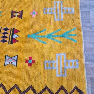 May include: Mustard yellow rug with geometric patterns. The rug features black zigzag borders, along with teal, pink, and gray shapes. The design includes cross-like figures and arrow-like motifs, creating a vibrant and textured look. The rug is made of a thick, woven material.