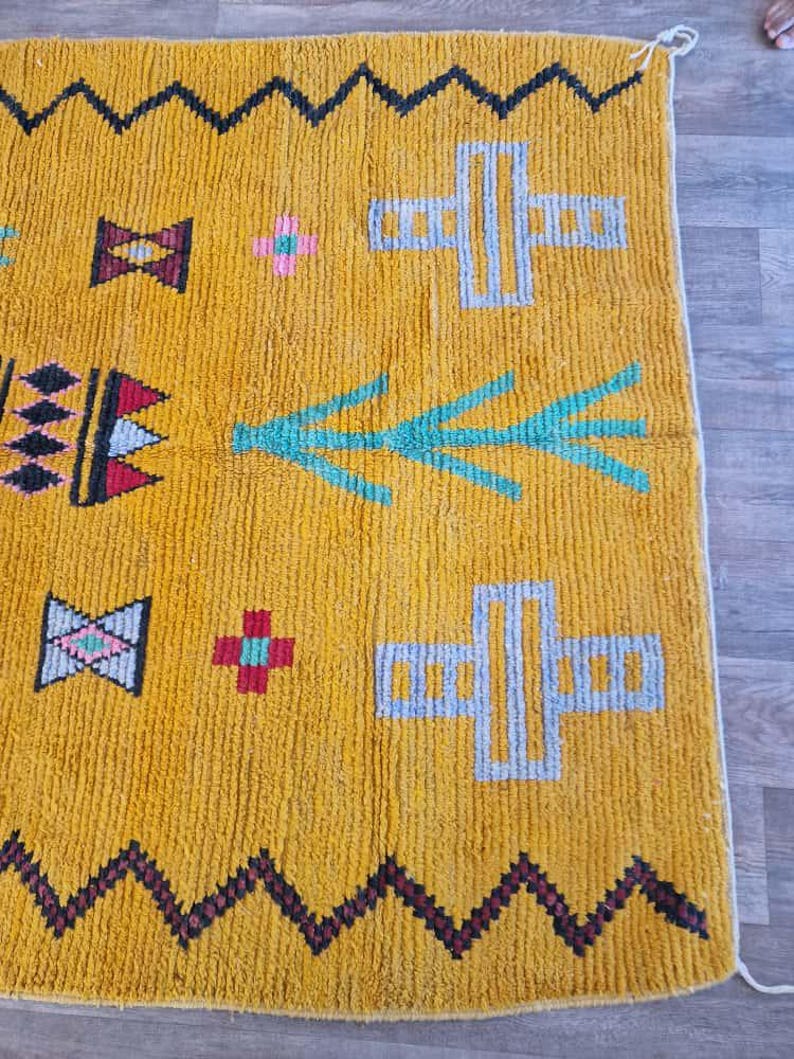 May include: Mustard yellow rug with geometric patterns. The rug features black zigzag borders, along with teal, pink, and gray shapes. The design includes cross-like figures and arrow-like motifs, creating a vibrant and textured look. The rug is made of a thick, woven material.