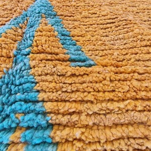 May include: Close-up of a textured rug with a geometric design. The rug features a combination of mustard yellow and turquoise blue colors. The design includes a triangle shape in the center and other shapes in the corners.
