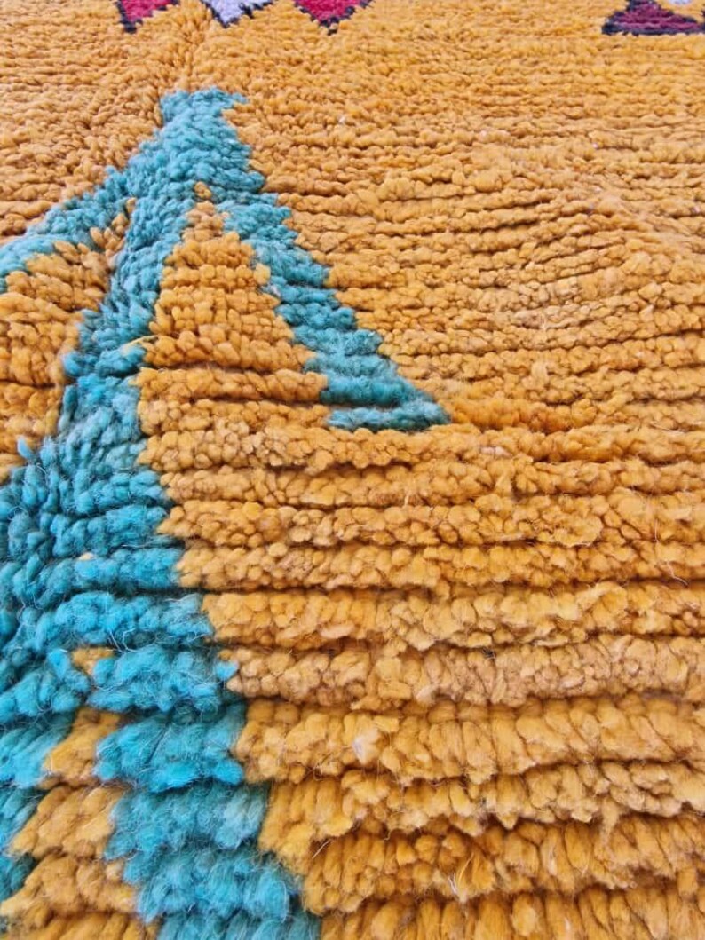 May include: Close-up of a textured rug with a geometric design. The rug features a combination of mustard yellow and turquoise blue colors. The design includes a triangle shape in the center and other shapes in the corners.