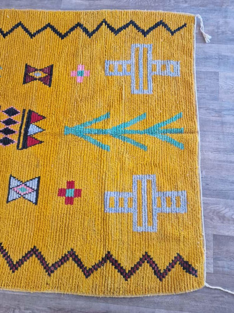 May include: Mustard yellow rug with geometric patterns. The rug features black zigzag borders, teal and gray cross shapes, and various other colorful geometric designs. The rug has a textured appearance and a neutral background.