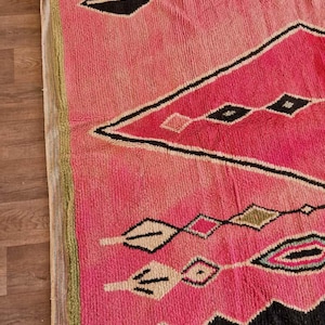 May include: A pink and black geometric patterned rug. The rug features a diamond and zigzag design with accents of white, green, and black. The rug is made of a thick, woven material.