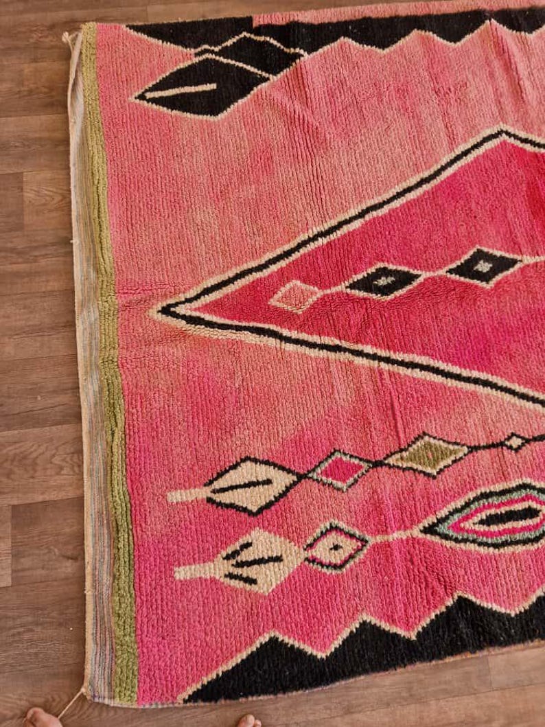 May include: A pink and black geometric patterned rug. The rug features a diamond and zigzag design with accents of white, green, and black. The rug is made of a thick, woven material.