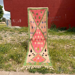 May include: A rectangular, hand-woven rug with a pink and green geometric design. The rug features diamond and triangle patterns in pink, green, and black, with a cream-colored background. The rug has a fringe along the bottom edge.
