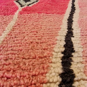 May include: Close-up of a pink and white Moroccan rug with a geometric design. The rug features a diamond pattern in the center, a black stripe, and a textured surface. The rug's color palette includes shades of pink, white, and black.