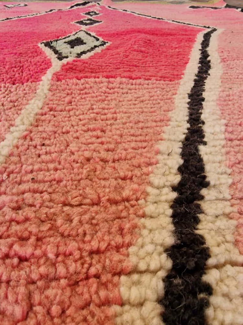 May include: Close-up of a pink and white Moroccan rug with a geometric design. The rug features a diamond pattern in the center, a black stripe, and a textured surface. The rug's color palette includes shades of pink, white, and black.