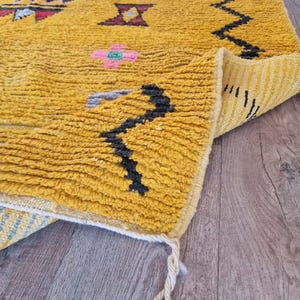 May include: Mustard yellow Moroccan rug with geometric patterns in black, pink, and teal. The rug has a textured, woven appearance and a folded corner revealing a striped underside. Tassels hang from the corner.