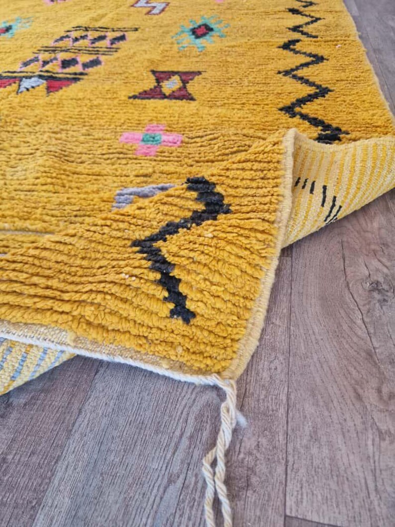 May include: Mustard yellow Moroccan rug with geometric patterns in black, pink, and teal. The rug has a textured, woven appearance and a folded corner revealing a striped underside. Tassels hang from the corner.