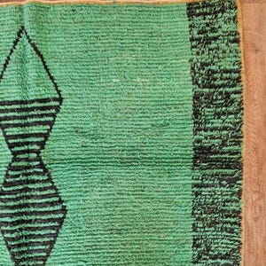 May include: A section of a green rug with a geometric design. The rug features a repeating pattern of black diamond shapes and a black border on the right side. The rug is made of a textured material.