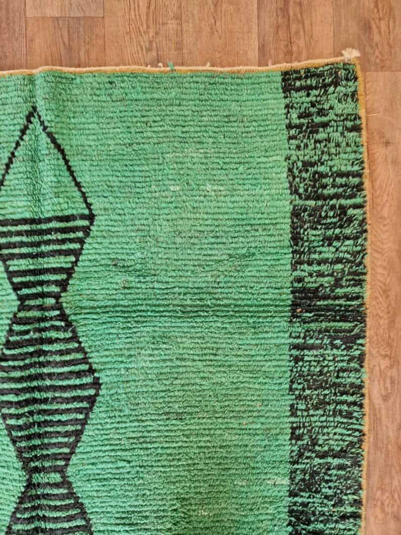 May include: A section of a green rug with a geometric design. The rug features a repeating pattern of black diamond shapes and a black border on the right side. The rug is made of a textured material.