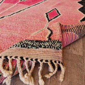 May include: A pink Moroccan rug with a geometric design featuring diamonds and other shapes in shades of pink and black. The rug has a thick pile and a fringe along one edge. The rug is partially rolled up, revealing the underside with colorful stripes.