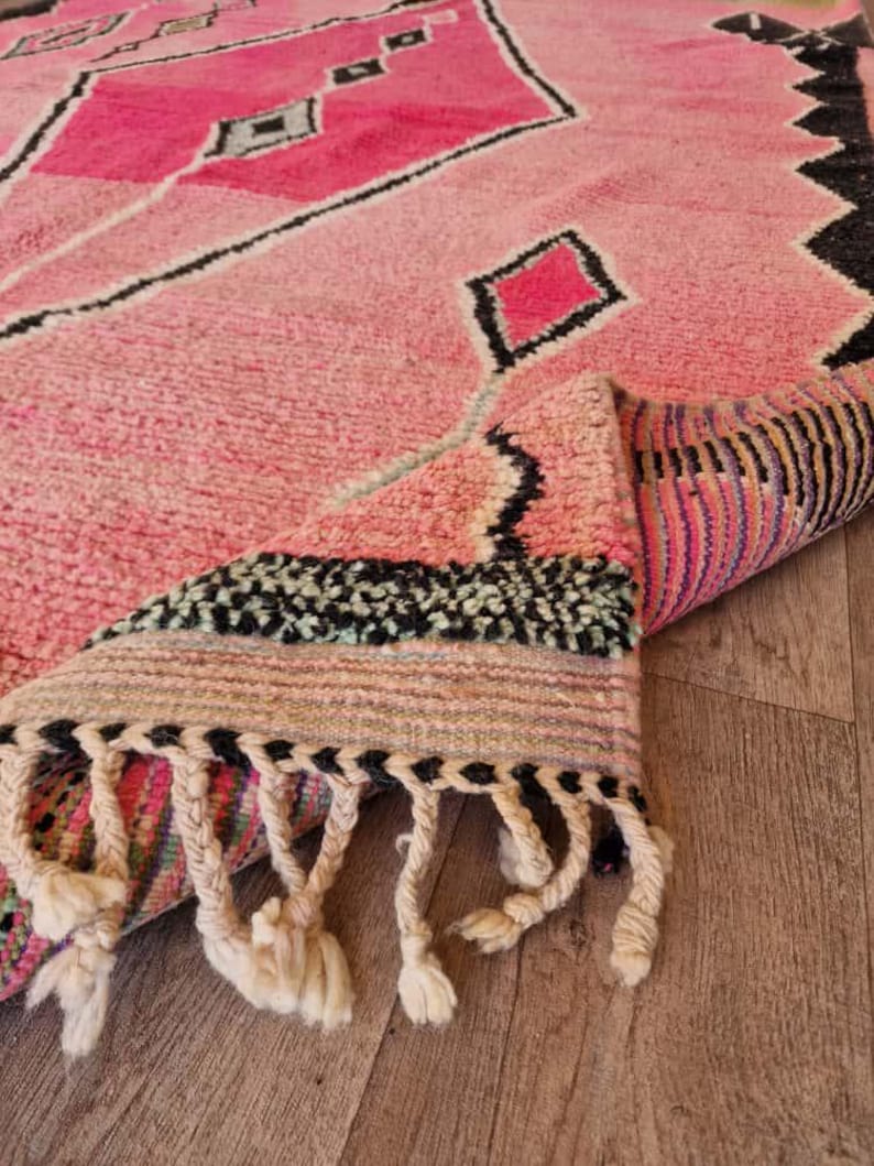 May include: A pink Moroccan rug with a geometric design featuring diamonds and other shapes in shades of pink and black. The rug has a thick pile and a fringe along one edge. The rug is partially rolled up, revealing the underside with colorful stripes.