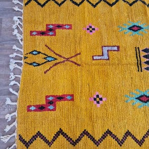 May include: Mustard yellow rug with geometric patterns in black, red, and turquoise. The rug features a zig-zag border and decorative tassels on one edge. The design includes various shapes and symbols.