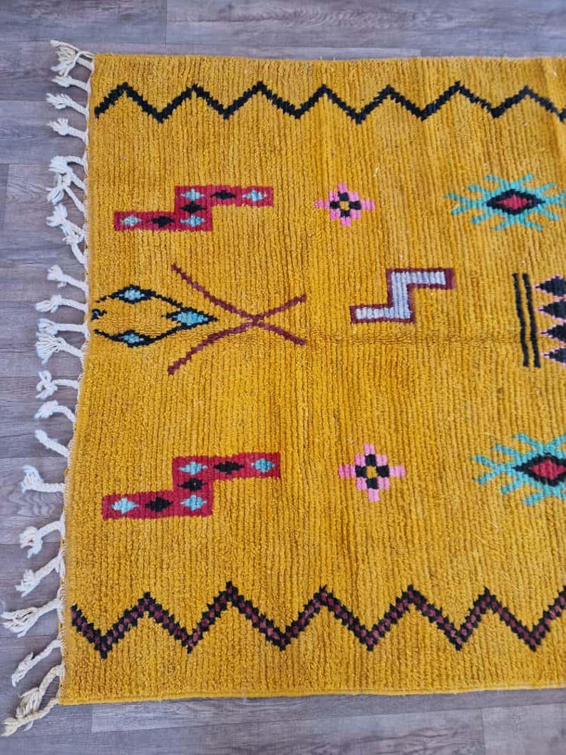 May include: Mustard yellow rug with geometric patterns in black, red, and turquoise. The rug features a zig-zag border and decorative tassels on one edge. The design includes various shapes and symbols.