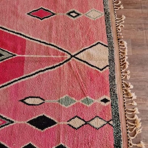 May include: A pink and black geometric patterned rug with diamond and zigzag designs. The rug has a fringe on one side and is made of a woven material. The rug's color palette includes shades of pink, black, white, and green.