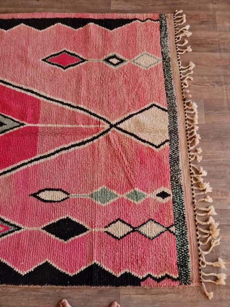 May include: A pink and black geometric patterned rug with diamond and zigzag designs. The rug has a fringe on one side and is made of a woven material. The rug's color palette includes shades of pink, black, white, and green.