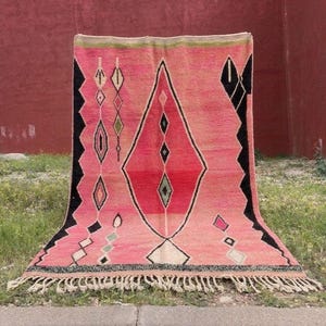 May include: A pink and red geometric rug with a diamond pattern. The rug has black and white accents, and a fringe along the bottom edge. The rug is displayed outdoors on grass.
