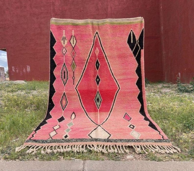 May include: A pink and red geometric rug with a diamond pattern. The rug has black and white accents, and a fringe along the bottom edge. The rug is displayed outdoors on grass.