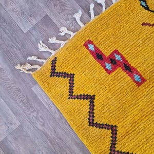May include: Mustard yellow rug with geometric patterns in black, red, and turquoise. The rug has a textured surface and white fringe along one edge. The design features a zig-zag pattern and other shapes.