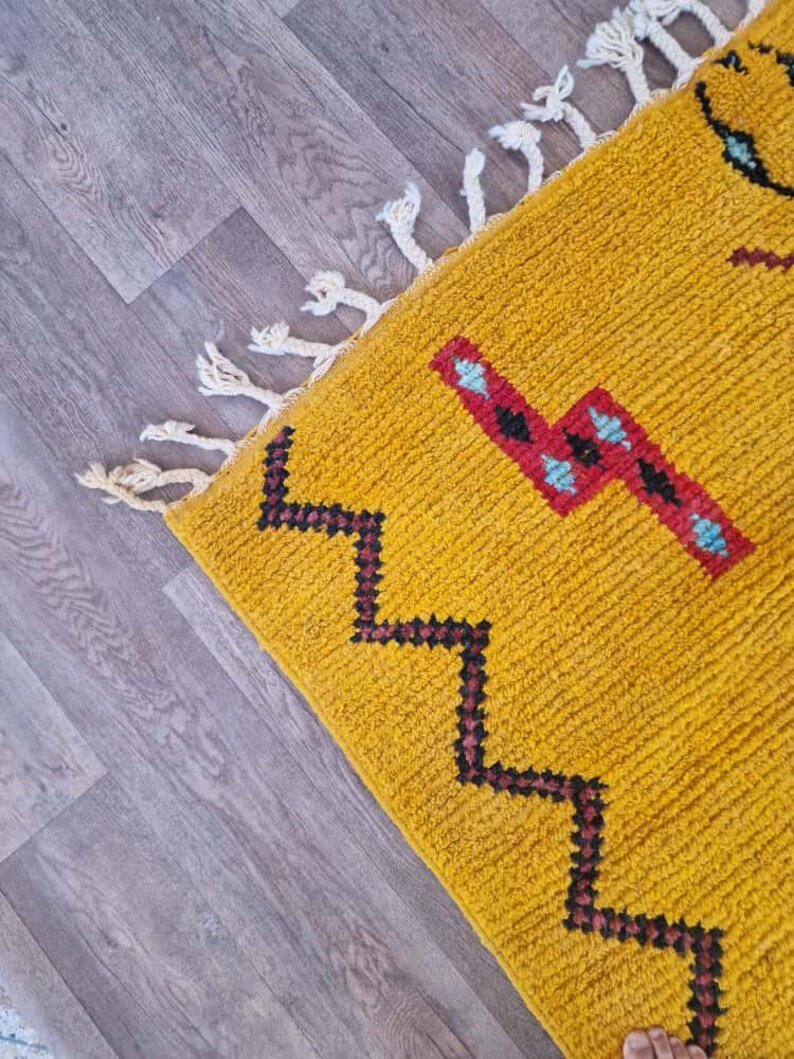 May include: Mustard yellow rug with geometric patterns in black, red, and turquoise. The rug has a textured surface and white fringe along one edge. The design features a zig-zag pattern and other shapes.