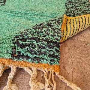 May include: A close-up of a hand-woven rug with a teal and black geometric pattern. The rug has a yellow and orange border with cream-colored tassels. The rug is made of a thick, textured material.