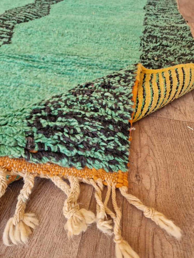 May include: A close-up of a hand-woven rug with a teal and black geometric pattern. The rug has a yellow and orange border with cream-colored tassels. The rug is made of a thick, textured material.