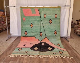 May include: A hand-woven rug with a geometric design. The rug features a light green background with pink and black shapes, and orange diamond accents. The rug has a fringe along the bottom edge.