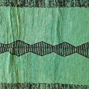 May include: A green and black rug with a textured surface. The rug features a central green field with a black zigzag pattern and black borders at the top and bottom. The rug is made of a thick, woven material.