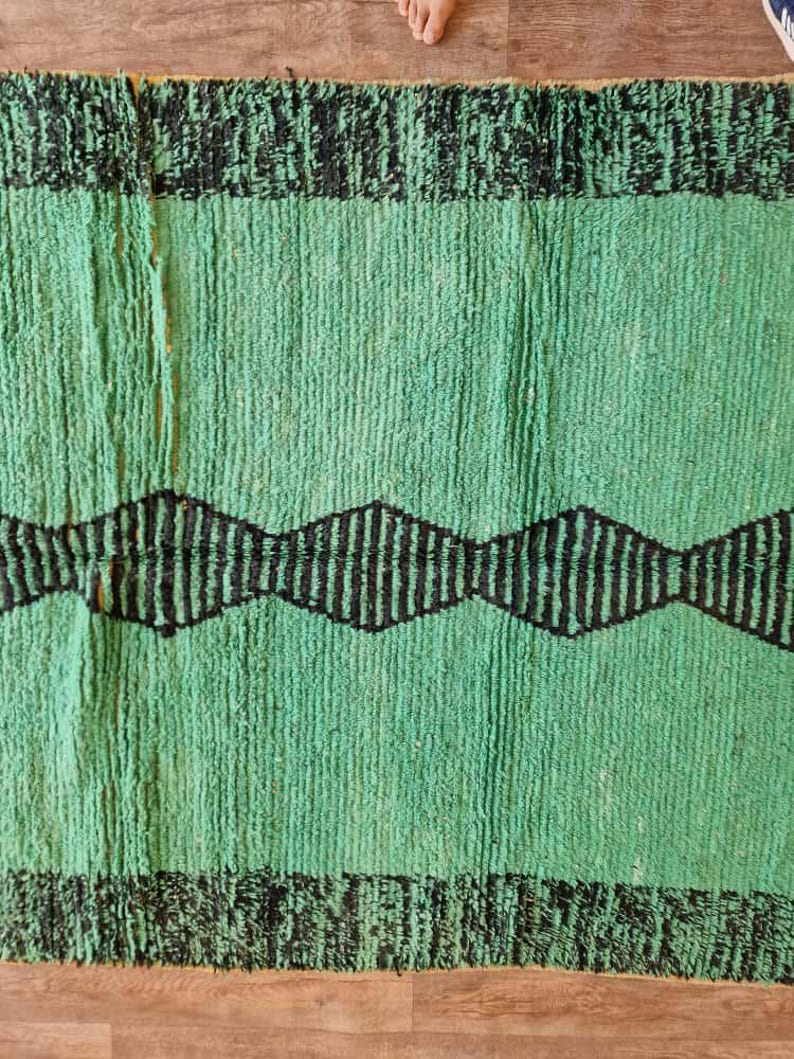 May include: A green and black rug with a textured surface. The rug features a central green field with a black zigzag pattern and black borders at the top and bottom. The rug is made of a thick, woven material.