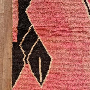 May include: Close-up of a pink and black geometric patterned rug. The rug features a textured, woven design with a gradient of pink hues and black diamond shapes. The rug has a green border.