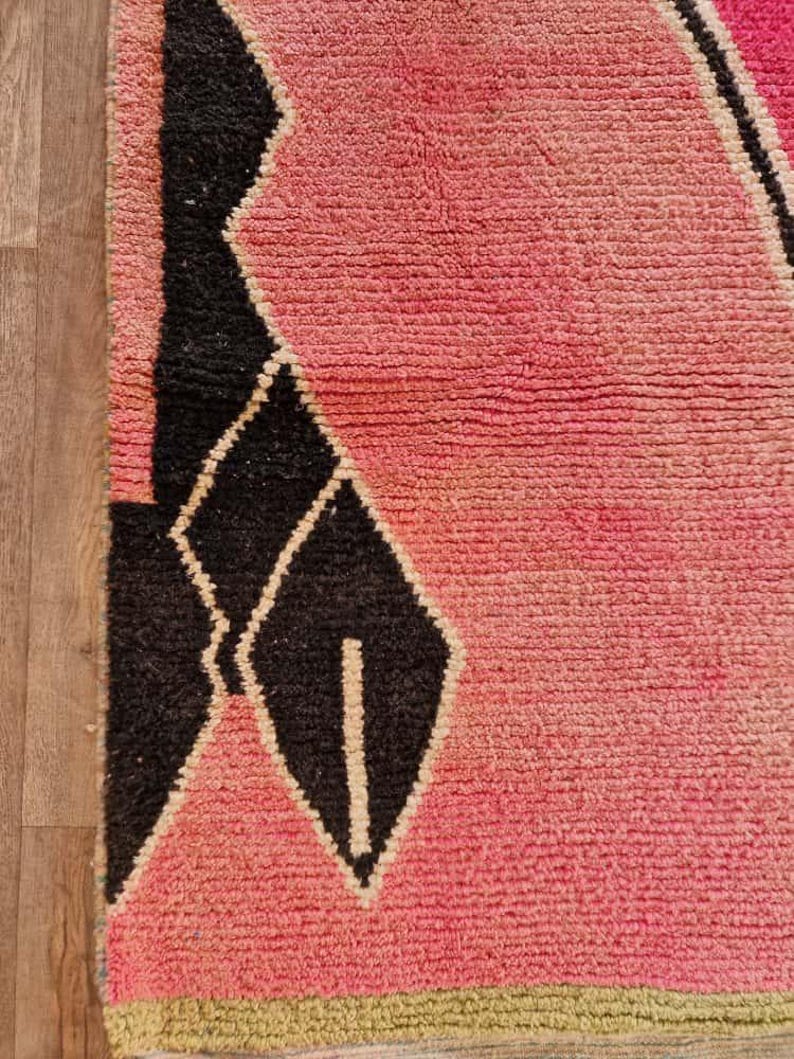 May include: Close-up of a pink and black geometric patterned rug. The rug features a textured, woven design with a gradient of pink hues and black diamond shapes. The rug has a green border.