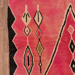 May include: A close-up of a vibrant pink rug with geometric patterns. The rug features diamond and arrow-like shapes in black, white, and various shades of pink and green. The rug's texture is visible, suggesting a woven or knotted construction.