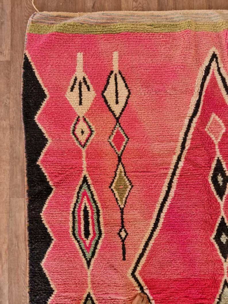 May include: A close-up of a vibrant pink rug with geometric patterns. The rug features diamond and arrow-like shapes in black, white, and various shades of pink and green. The rug's texture is visible, suggesting a woven or knotted construction.